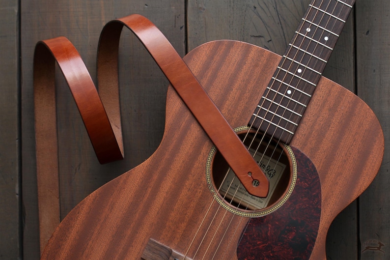 Puede incluir: Guitarra ac&uacute;stica con una correa de cuero marr&oacute;n. La guitarra tiene un cuerpo de madera con un golpeador marr&oacute;n oscuro y una boca. La correa est&aacute; enganchada a la guitarra y es de un color marr&oacute;n c&aacute;lido y rico.