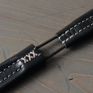 May include: Close-up of a black leather camera strap with white stitching. The strap is adjustable and has a simple design.