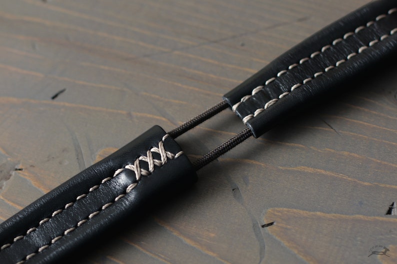 May include: Close-up of a black leather camera strap with white stitching. The strap is adjustable and has a simple design.