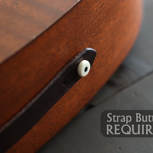 Op de afbeelding: Close-up van een bruine houten gitaar met een zwarte leren riem en een witte riemknop. De tekst "Strap Button REQUIRED" is zichtbaar onder de gitaar.