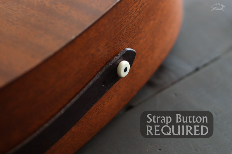Op de afbeelding: Close-up van een bruine houten gitaar met een zwarte leren riem en een witte riemknop. De tekst "Strap Button REQUIRED" is zichtbaar onder de gitaar.