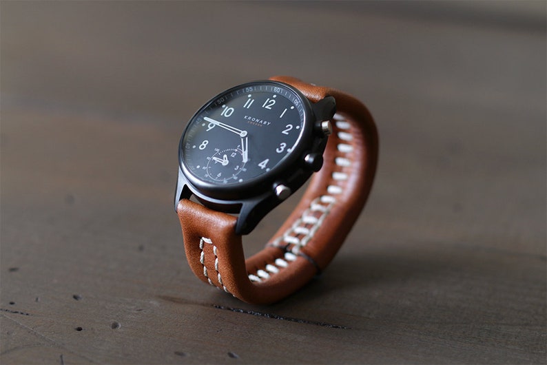 May include: A black and silver smartwatch with a brown leather band. The watch face has a black background with white numbers and hands. The watch is on a wooden surface.