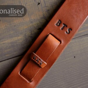 May include: A close-up of a brown leather guitar strap with the initials "BTS" embossed on it. The strap has a buckle and a loop for adjustment. The text "personalised custom text" is in the upper left corner.