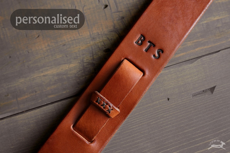 May include: A close-up of a brown leather guitar strap with the initials "BTS" embossed on it. The strap has a buckle and a loop for adjustment. The text "personalised custom text" is in the upper left corner.