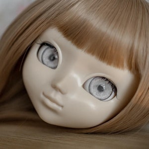 Puede incluir: Una cabeza de muñeca con cabello largo, liso y castaño claro y ojos grandes y blancos. La muñeca tiene una sonrisa pintada y mira directamente a la cámara.