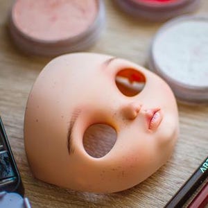 May include: A doll head with open eye sockets and painted lips rests on a wooden surface. Makeup supplies, including powder compacts, a watercolor palette, and Arteza pencils, are arranged around the doll head, suggesting a crafting or art project.