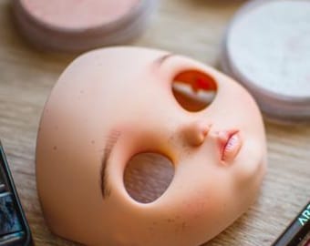 May include: A doll head with open eye sockets and painted lips rests on a wooden surface. Makeup supplies, including powder compacts, a watercolor palette, and Arteza pencils, are arranged around the doll head, suggesting a crafting or art project.