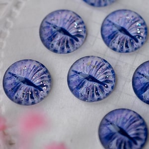 May include: A collection of ten round, glass eye cabochons. Each cabochon features a detailed, blue iris with a dark pupil and white highlights, set against a lighter blue background. The eyes are arranged on a white, lacy surface.