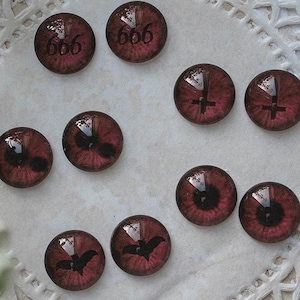 May include: A selection of small, circular, red-hued glass cabochons with different motifs. Some have the number "666", others a cross, a bat, or an eye. They are displayed on a white, lace-like surface, with green foliage in the background.