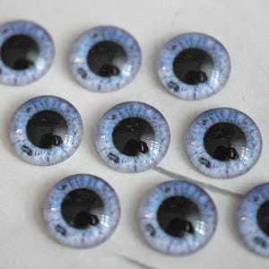 Op de afbeelding: Negen glazen poppenogen met een lichtblauwe iris en een zwarte pupil liggen op een wit, wolkvormig dienblad. De ogen hebben een glanzende afwerking en zijn geschikt voor poppenmaken of knutselen.