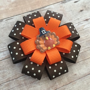 May include: A brown and white polka dot ribbon bow with an orange ribbon bow in the center. A small turkey with a pilgrim hat is in the center of the orange bow.