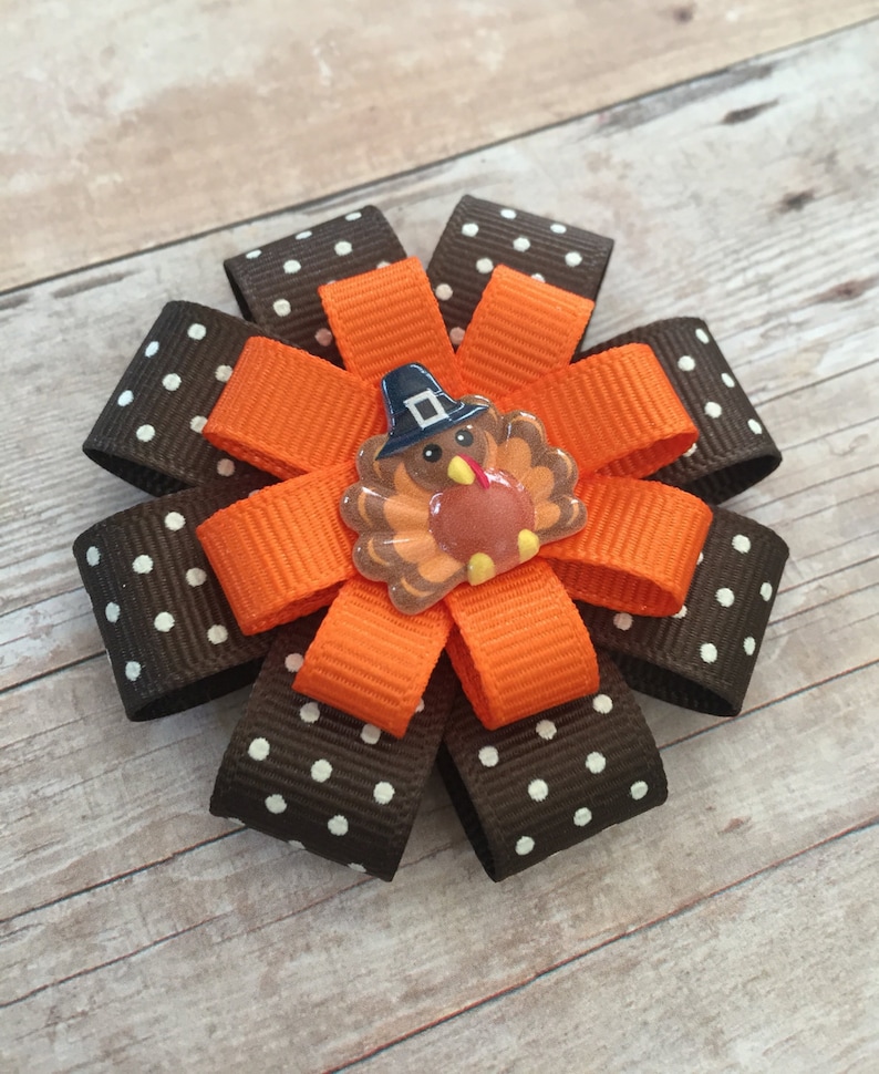 May include: A brown and white polka dot ribbon bow with an orange ribbon bow in the center. A small turkey with a pilgrim hat is in the center of the orange bow.