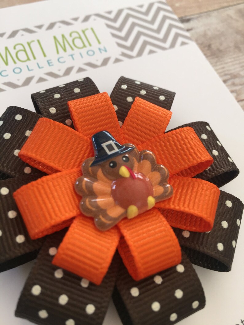 May include: A brown and white polka dot ribbon bow with an orange ribbon and a turkey-shaped embellishment. The turkey is wearing a blue hat and has a red beak and orange feet.