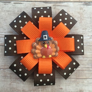 May include: A brown and white polka dot ribbon bow with an orange ribbon bow in the center. A small turkey with a pilgrim hat is in the center of the orange bow.