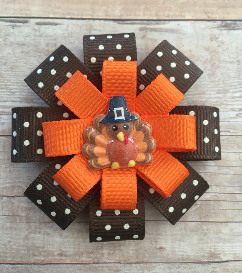 May include: A brown and white polka dot ribbon bow with an orange ribbon bow in the center. A small turkey with a pilgrim hat is in the center of the orange bow.