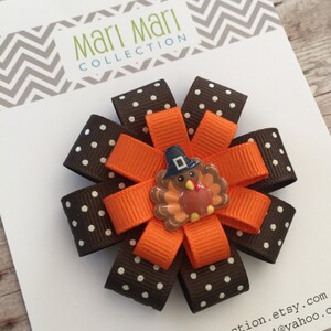 May include: A brown and white polka dot ribbon bow with an orange ribbon and a Thanksgiving turkey applique. The bow is attached to a white card with the text "Mari Mari Collection" and contact information.