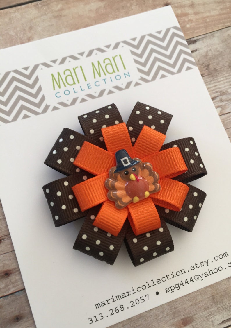 May include: A brown and white polka dot ribbon bow with an orange ribbon and a Thanksgiving turkey applique. The bow is attached to a white card with the text "Mari Mari Collection" and contact information.