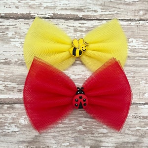 May include: Two tulle hair bows with decorative embellishments. The top bow is yellow with a black and yellow bee. The bottom bow is red with a black and red ladybug.