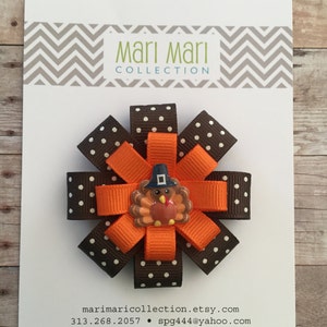 May include: A brown and orange hair bow with white polka dots and a turkey charm. The bow is made of ribbon and has a clip on the back. The charm is a small, round turkey with a brown body, orange legs, and a red wattle. The turkey is wearing a blue hat.