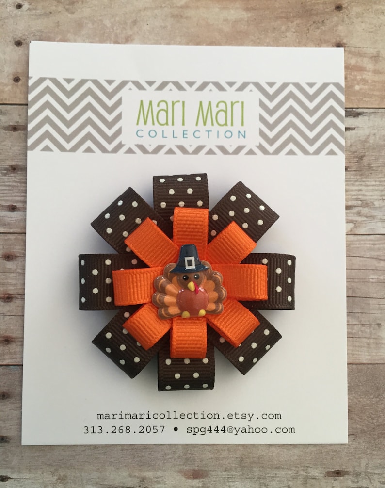 May include: A brown and orange hair bow with white polka dots and a turkey charm. The bow is made of ribbon and has a clip on the back. The charm is a small, round turkey with a brown body, orange legs, and a red wattle. The turkey is wearing a blue hat.