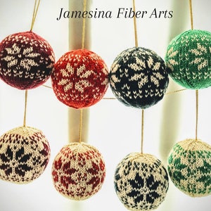 May include: Eight knitted Christmas ornaments in various colors, including red, green, and navy blue, each with a white snowflake pattern. The ornaments are suspended by gold strings. The text "Jamesina Fiber Arts" is in the upper left corner.