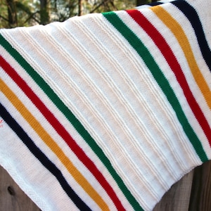 May include: White knitted throw blanket with colorful stripes in red, green, yellow, and navy blue. The blanket is folded and draped over a wooden fence. The text "Jamesina Fiber Art" is visible in the lower left corner.