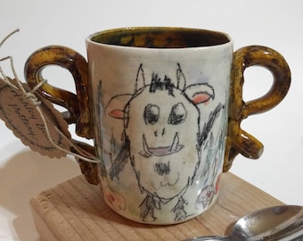 May include: A ceramic mug with two brown handles and a hand-drawn illustration of a monster with a toothy grin. The mug is sitting on a wooden surface with a silver spoon.