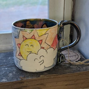 Pode incluir: Caneca de cerâmica pintada à mão com um design de sol e nuvem sorridentes. A caneca tem uma alça preta e um interior colorido. O design exterior inclui detalhes em amarelo, vermelho e azul, criando uma estética alegre. Uma peça única para desfrutar de bebidas.