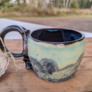 Puede incluir: Taza de cerámica hecha a mano con un diseño único. La taza presenta un exterior azul claro con detalles en azul oscuro y un asa negra. El interior es azul oscuro. La taza está sobre una superficie de madera.