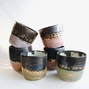 Può includere: Sei tazze in ceramica con smalto nero e dettagli dorati. Le tazze hanno interni di diversi colori, tra cui rosa, verde e beige.