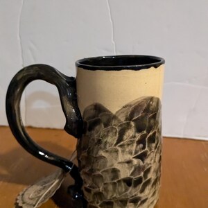 Puede incluir: Una taza de cerámica con asa negra y un diseño marrón y beige. El diseño se asemeja a un patrón texturizado de escamas.