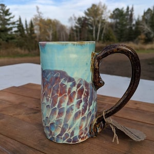 May include: A ceramic mug with a brown handle and a blue, green, and brown glaze. The mug has a unique, textured surface.