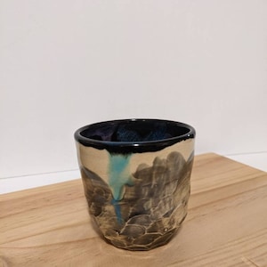 Puede incluir: Una taza de cerámica con un esmalte texturizado marrón y negro. La taza tiene un diseño azul oscuro y blanco en el lateral.