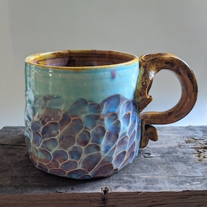 Puede incluir: Una taza de cerámica hecha a mano con un esmalte azul y morado y un diseño texturizado y orgánico. La taza tiene una asa grande de color marrón.