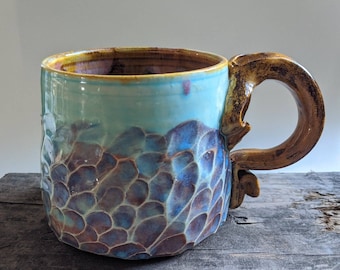 Puede incluir: Una taza de cerámica hecha a mano con un esmalte azul y morado y un diseño texturizado y orgánico. La taza tiene una asa grande de color marrón.