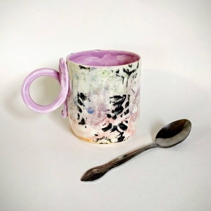 May include: A ceramic mug with a unique, abstract design in shades of pink, white, and black. The mug has a purple handle that is shaped like a loop. A silver spoon is lying on the table next to the mug.