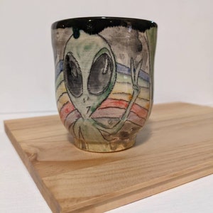 May include: A ceramic mug with a black rim and a hand-painted design of a green alien with a rainbow background. The alien is making a peace sign with its hand.