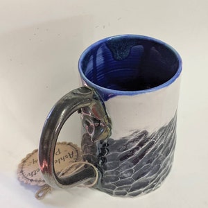 Puede incluir: Una taza de cerámica blanca con un esmalte azul en el interior y un esmalte blanco y negro en el exterior. La taza tiene un asa única con forma de cabeza de dragón. La taza está sobre una superficie blanca. La taza tiene una etiqueta adjunta al asa que dice "Ashlie p."