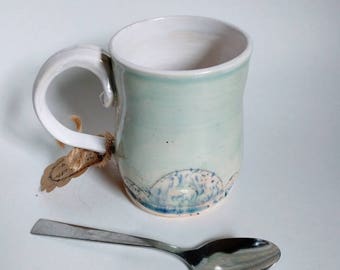 May include: A light blue ceramic mug with a white interior and a blue and white wave design around the bottom. The mug has a handle and is sitting on a white surface with a silver spoon next to it.