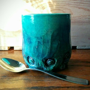 May include: A teal blue ceramic mug with a textured surface and raised floral designs. The mug is sitting on a wooden surface next to a silver spoon.