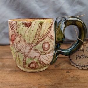 May include: A ceramic mug with a green glaze and a floral design. The mug has a unique handle and a tag that reads "Nicky Beth Pottery".