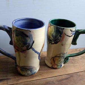 Op de afbeelding: Twee keramische mokken met abstracte bloemenmotieven. De ene mok heeft een blauwe rand en een blauwe handgreep, de andere heeft een groene rand en een groene handgreep.
