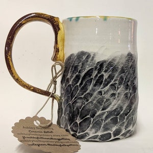 Puede incluir: Una taza de cerámica con un diseño texturizado en blanco y negro. La taza tiene un asa amarilla y un borde blanco. La taza está sobre una superficie blanca. La taza tiene una etiqueta adjunta que dice "Ashley Nordberg Ceramic Artist ashleybethpottery.etsy.com ashleybethnordbergpottery ashleybethnordberg@gmail.com instagram @ashleybethpottery"