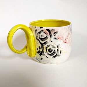 May include: A white ceramic mug with a yellow handle and a black and white geometric pattern. The mug has a yellow interior.