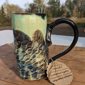 Puede incluir: Una taza de cerámica hecha a mano con un esmalte azul, verde y marrón. La taza tiene una superficie texturizada única y un asa negra. La taza está sobre una mesa de madera con una etiqueta de papel marrón unida al asa. La etiqueta tiene el texto "Norberg Ceramic Artist" y la información de contacto.