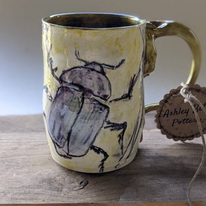 Peut inclure: Une tasse en céramique jaune et verte avec un motif de scarabée peint à la main. La tasse a une forme arrondie et une anse. La tasse est signée "Ashley Potter".