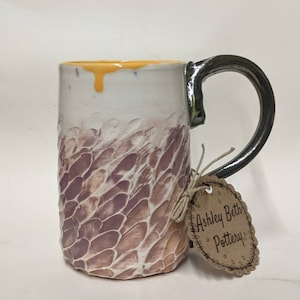 Puede incluir: Una taza de cerámica blanca con un diseño texturizado morado y marrón. La taza tiene un asa negra y un goteo de esmalte amarillo. La taza tiene una etiqueta que dice "Ashley Beth Pottery".