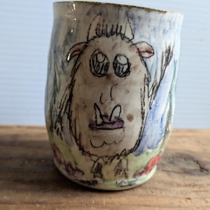 Puede incluir: Una taza de cerámica blanca con una ilustración pintada a mano de una criatura peluda con ojos grandes y un sombrero morado. El fondo es una acuarela azul y verde.
