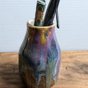 May include: A small, iridescent ceramic vase with a dark blue, purple, and gold glaze. The vase is filled with a black pen and a small, green and gold book.