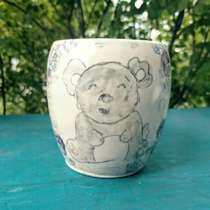 May include: A white ceramic mug with a hand-drawn illustration of a smiling bear with flowers around the rim.